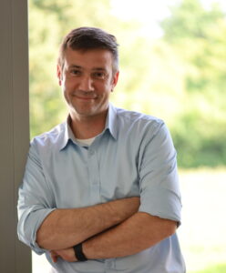 Mann mit kurz geschnittenem Haar in hellblauem Hemd vor unscharfem, grünem Naturhintergrund.