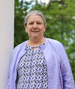 Ältere Frau mit grauerem Haar trägt lila gesteppte Jacke und gemustertes Kleid vor grünem Hintergrund.