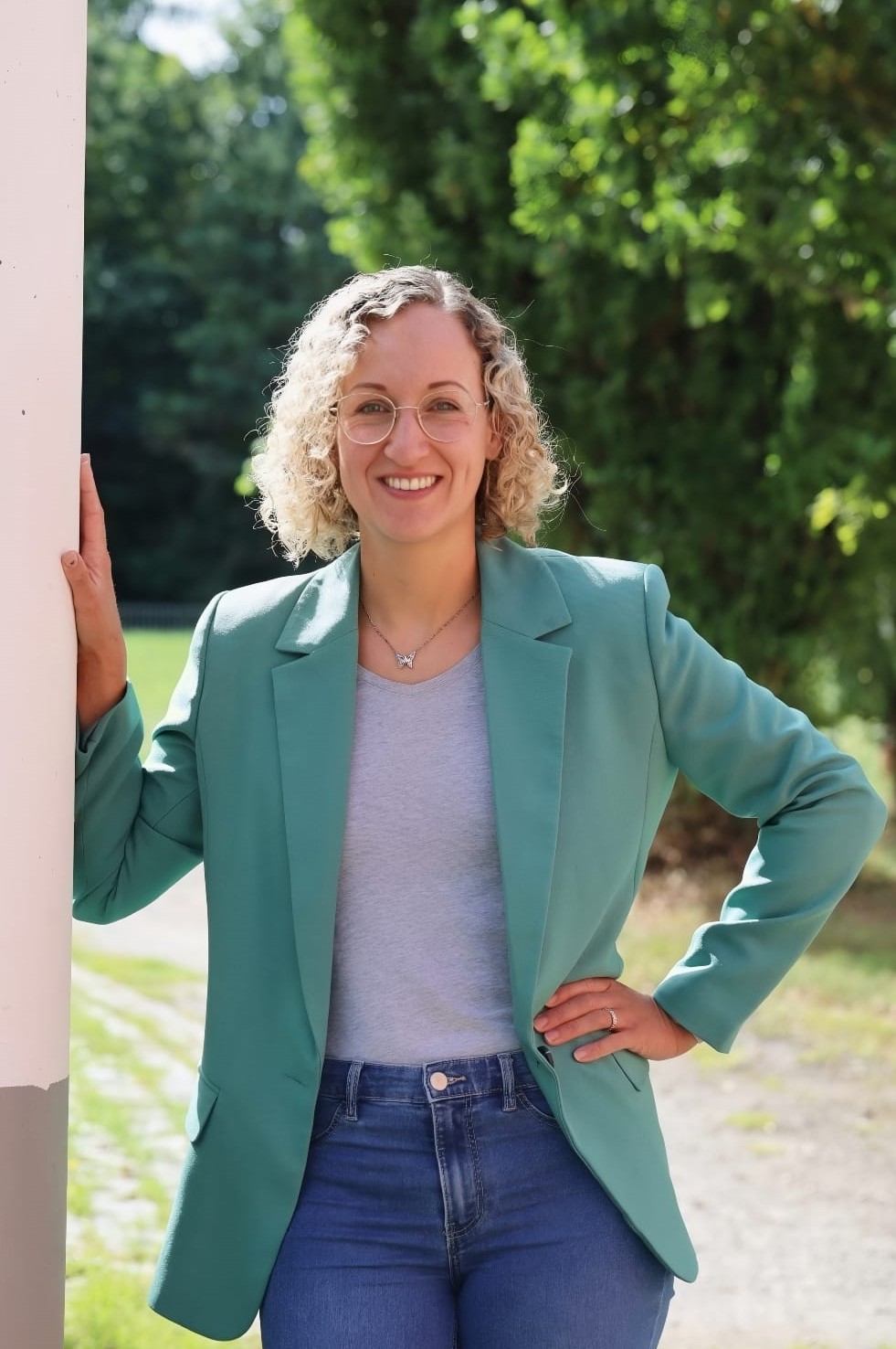 Frau mit lockigem blondem Haar, Brille, grünem Blazer und blauen Jeans lehnt an Wand im Freien.