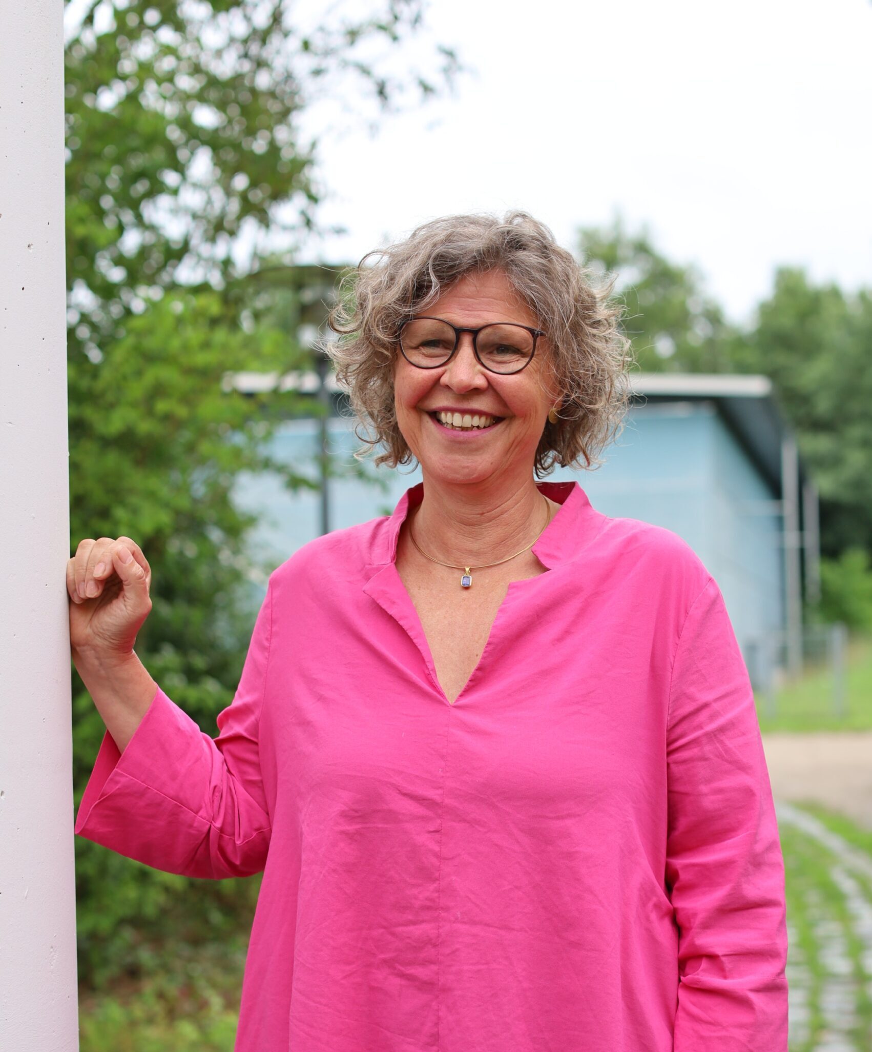 Frau mit kurzen lockigen Haaren und Brille in pinkfarbenem Oberteil lehnt an weißer Wand draußen.