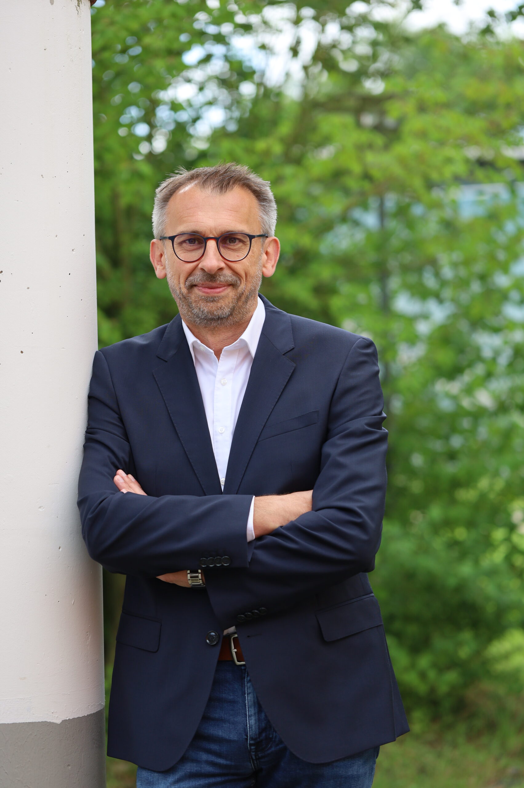 Mann mit Brille im dunkelblauen Anzug und weißem Hemd lehnt an weißer Säule vor grünem Laub.