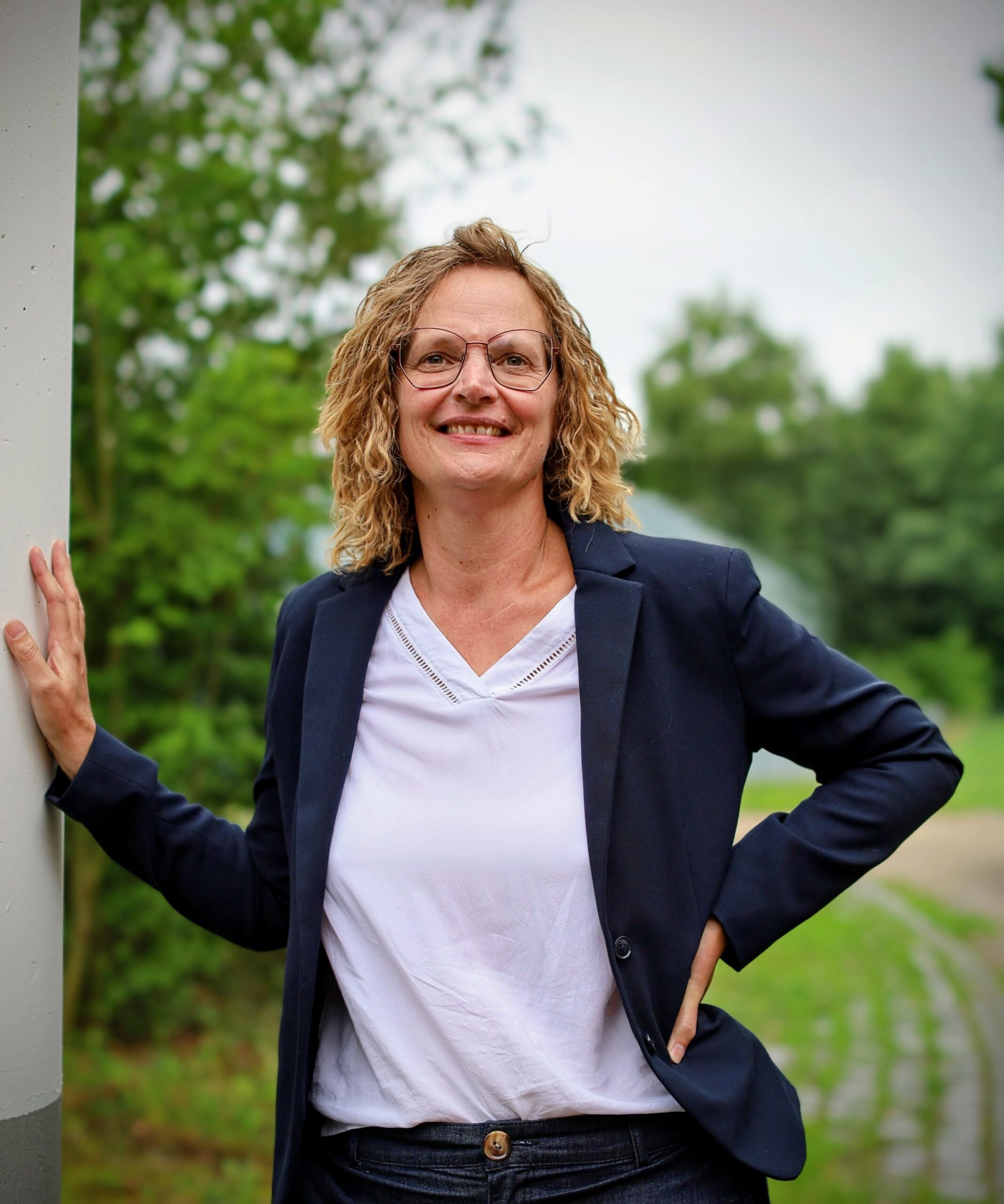 Frau mit lockigem Haar und Brille lehnt an Säule, trägt weißes Oberteil und blaues Sakko.