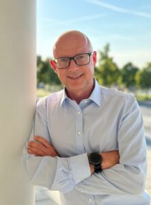 Mann mit Brille und hellblauem Hemd lehnt mit verschränkten Armen an weißer Säule im Freien.