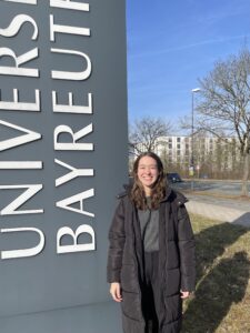 Frau steht vor Universität Bayreuth Schild mit Straße, Bäumen und Gebäude im Hintergrund.