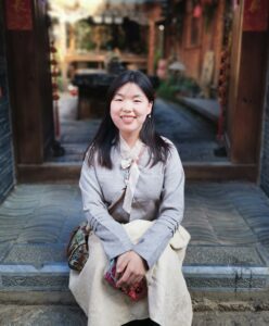 Frau sitzt vor traditionellem Holzgebäude auf Stufe, trägt grauen Pullover und hellen Rock.