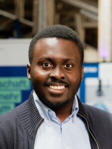 Porträt eines lächelnden Mannes mit blauem Hemd und dunkler Strickjacke vor unscharfem Hintergrund.