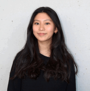 Junge Frau mit langen schwarzen Haaren und schwarzem Pullover vor heller Wand.