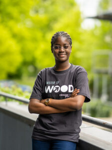 Junge Frau mit Brille und "MASTER OF WOOD" T-Shirt lehnt an Geländer vor grünem Hintergrund.