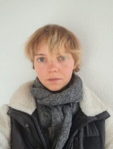 Person mit kurzen blonden Haaren, grauem Schal und schwarzer Jacke mit weißem Futter vor weißer Wand.