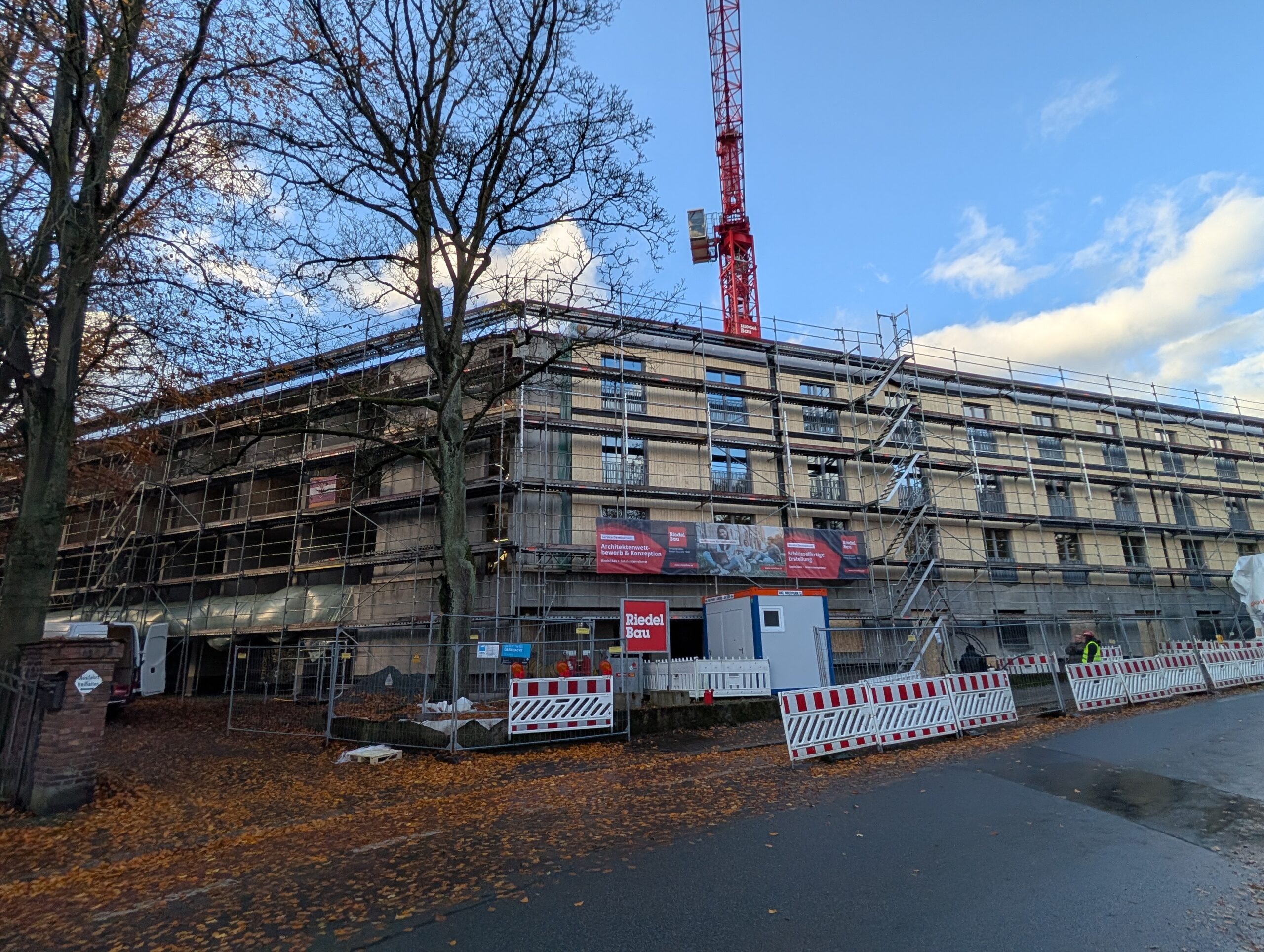 Mehrstöckiges Rohbaugebäude mit Gerüst, rotem Baukran, Baustellensicherungen und herbstlichen Bäumen.